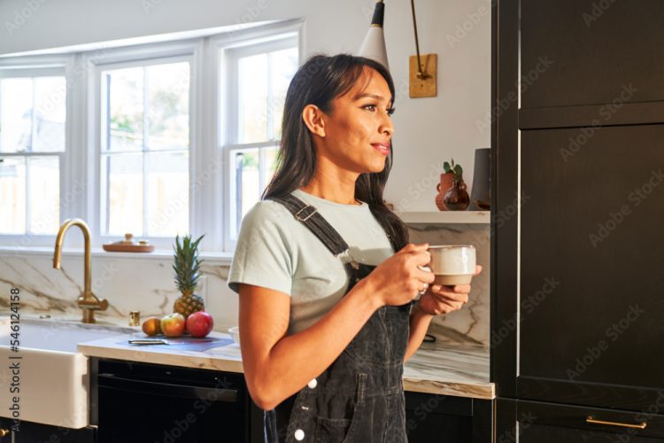 Frau mit langen dunklen Haaren steht in einer modernen Küche mit schwarzer und weißer Einrichtung, hält eine Kaffeetasse und blickt nachdenklich aus dem Fenster. Auf der Arbeitsplatte liegen frische Früchte und eine Ananas.
