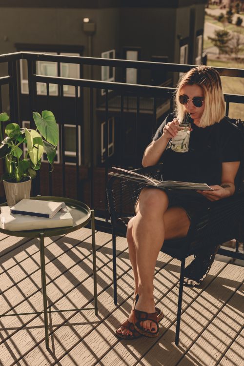 Frau in einem Sommer Outfit sitzt auf einem Balkon und trinkt aus einer Tasse, während sie ein Magazin liest.