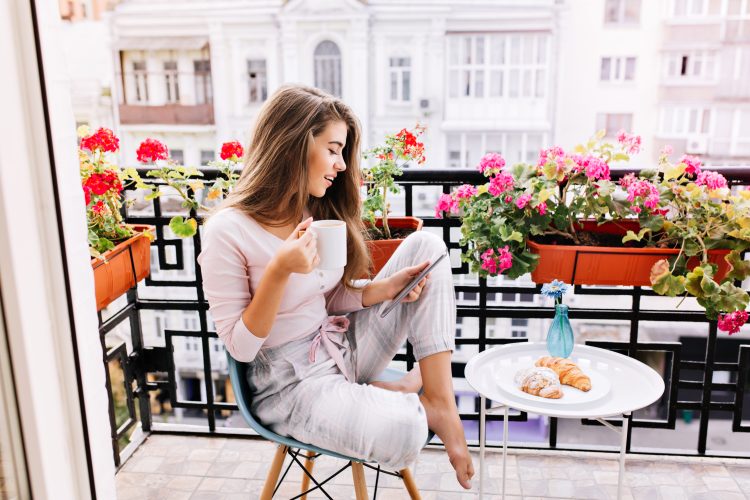 Attraktives Mädchen mit langen Haaren im Pyjama beim Frühstück auf dem Balkon am Morgen in der Stadt. Sie hält eine Tasse und liest auf dem Tablett.