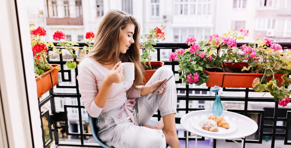 Attraktives Mädchen mit langen Haaren im Pyjama beim Frühstück auf dem Balkon am Morgen in der Stadt. Sie hält eine Tasse und liest auf dem Tablett.