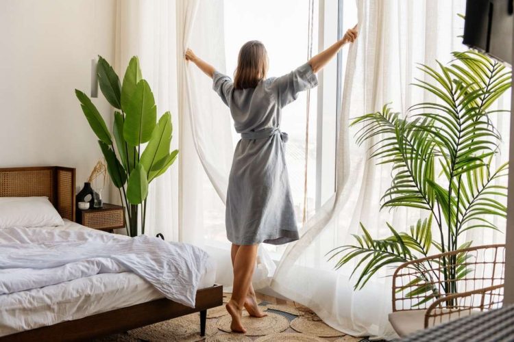 Frau macht Gardinen zur Seite und steht in ihrem Schlafzimmer
