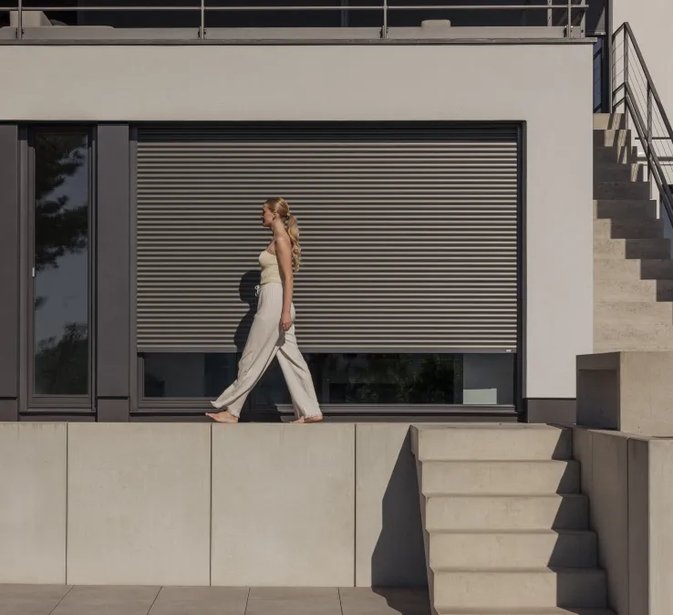 Frau läuft auf der Terrasse vor einem Fenster entlang - der Rolladen ist halb runtergelassen