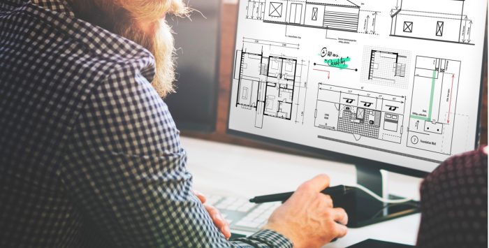 Mann mit Bart sitzt vor einem Computer und plant digital den Umriss eines Hauses.